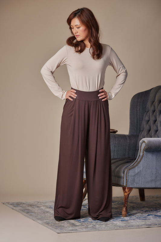 Woman wearing a beige top and brown wide-leg pants standing in a room with a blue armchair.