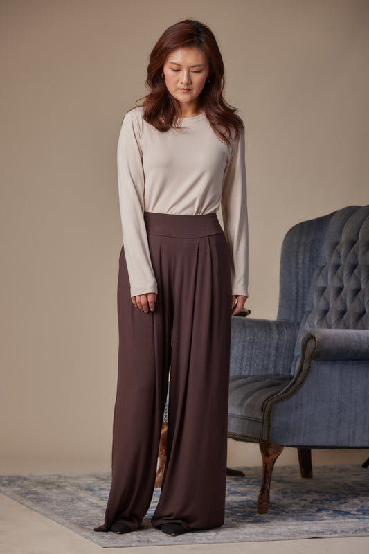 Woman wearing a beige top and brown wide-leg pants standing in a room with a blue armchair.