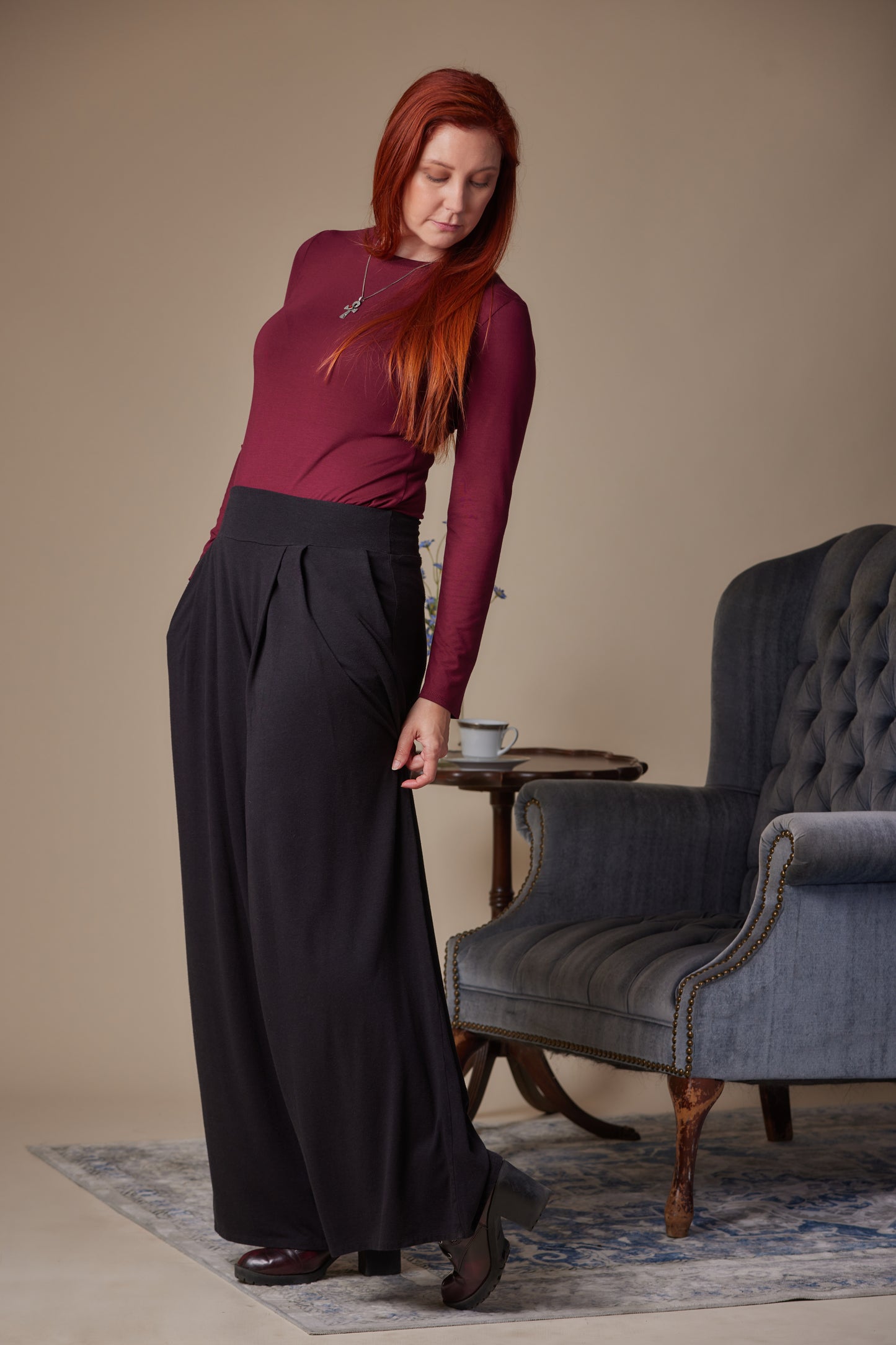 Woman in a burgundy top and black pants standing in a room with a gray armchair and small table.