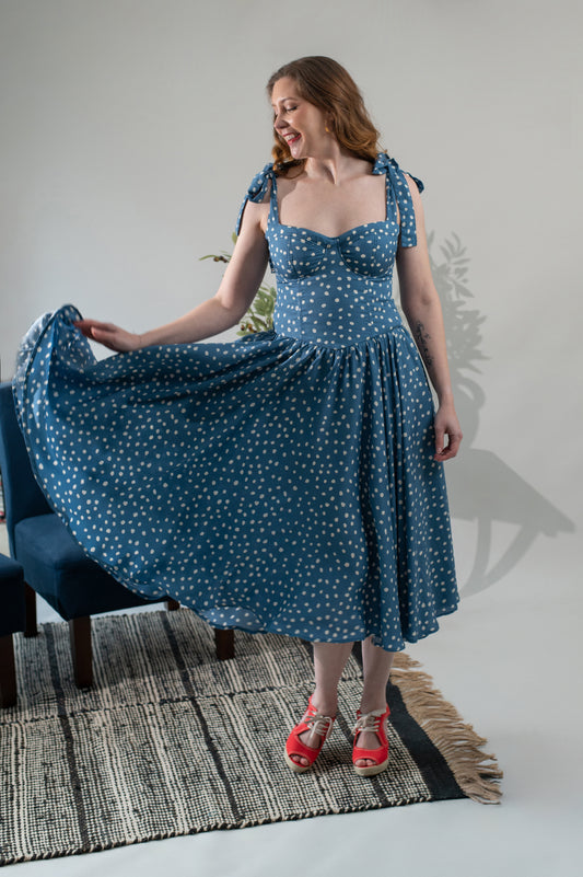 Woman in a blue polka dot dress standing in a room with a chair and rug.
