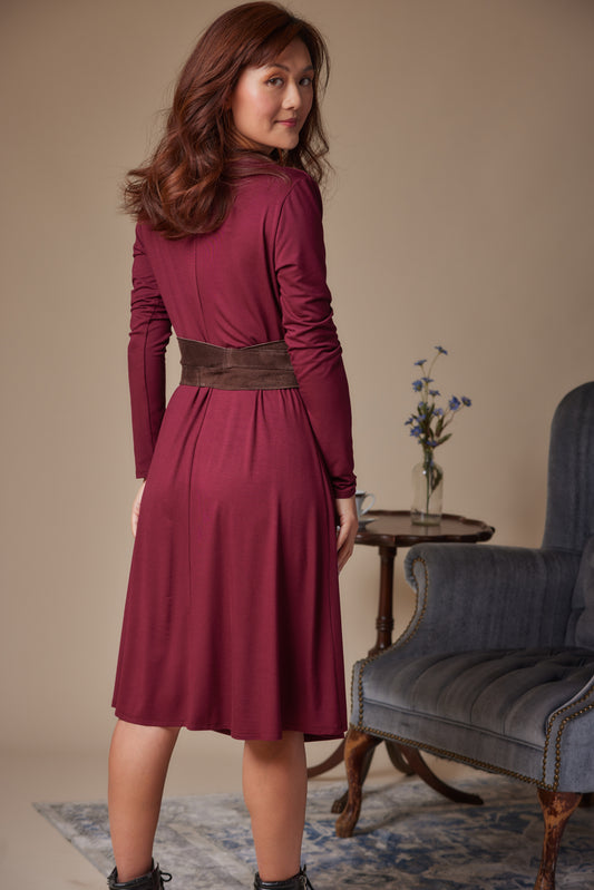Woman wearing a burgundy dress with a brown belt in a room with a blue armchair and small table.