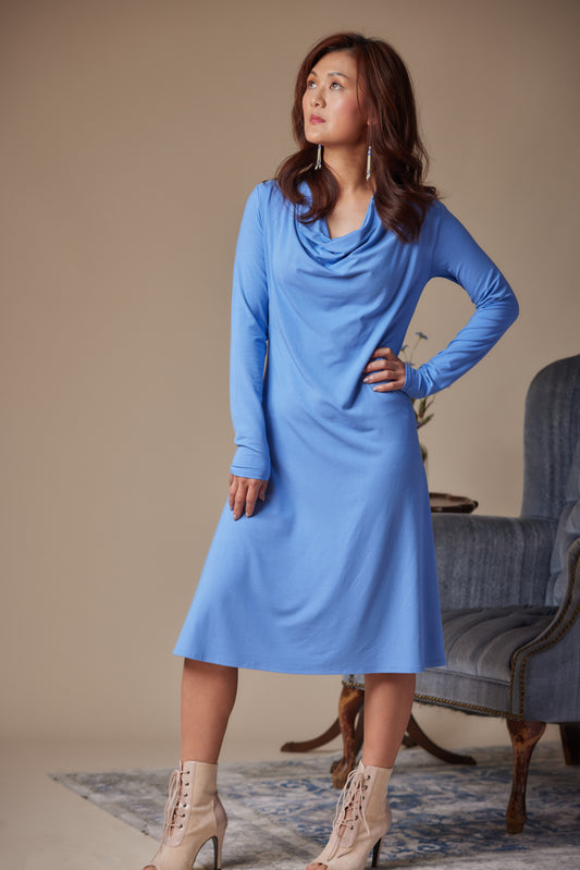 Woman wearing a blue dress standing in a room with a beige wall and a gray chair.