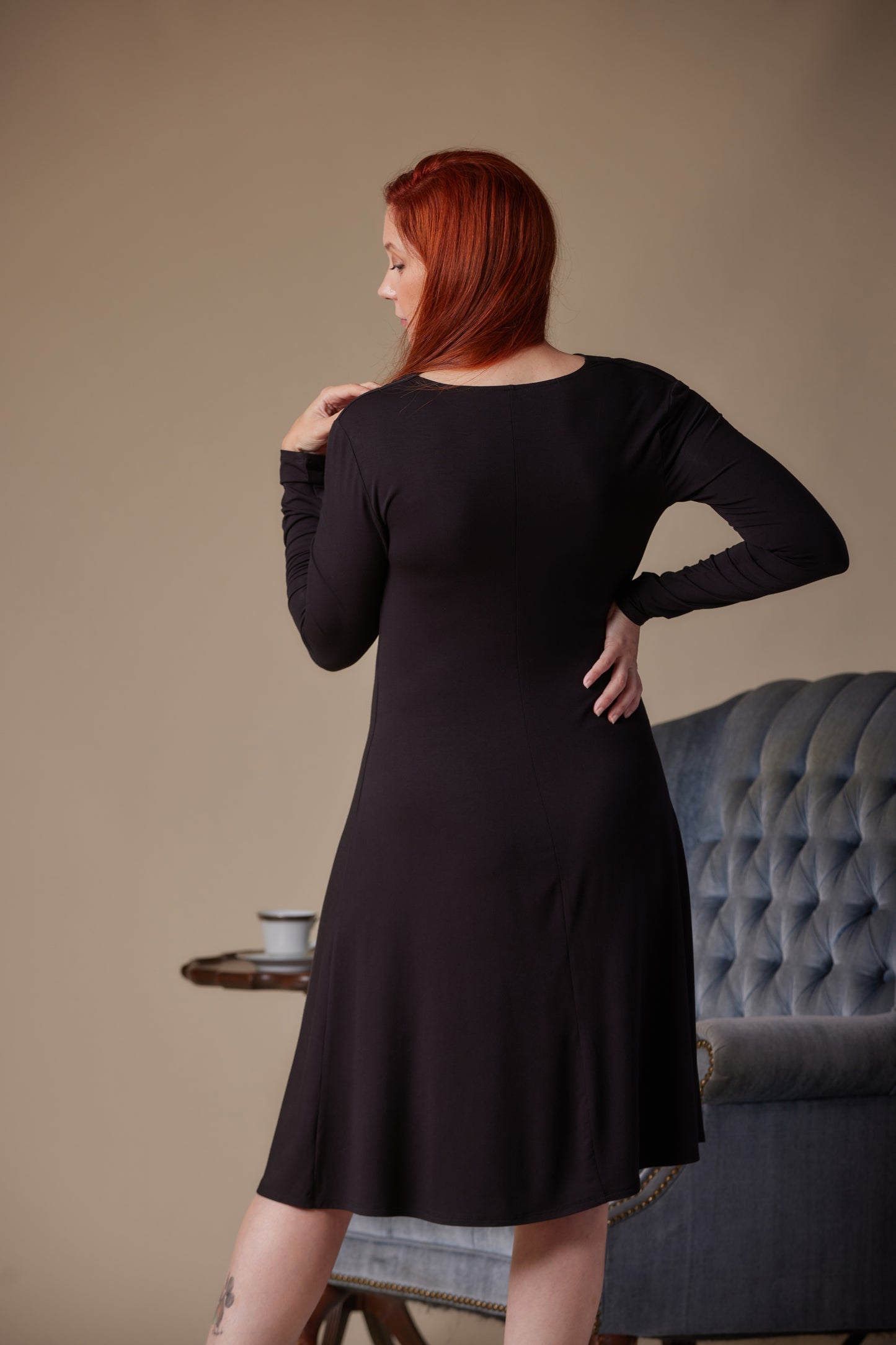Woman wearing a black dress standing in a room with a gray sofa and beige wall.