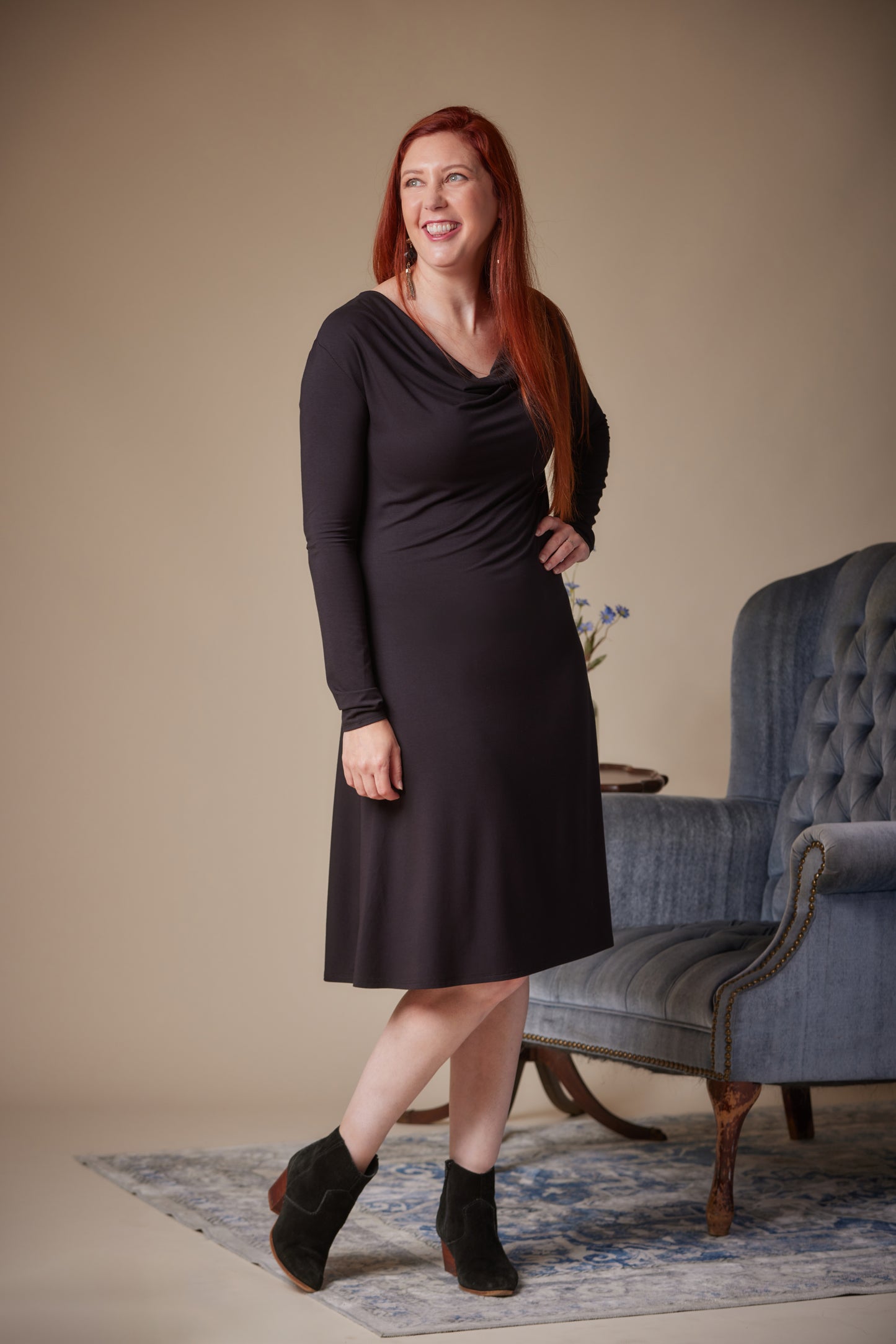 Woman in a black dress standing in a room with a blue armchair and floral arrangement.