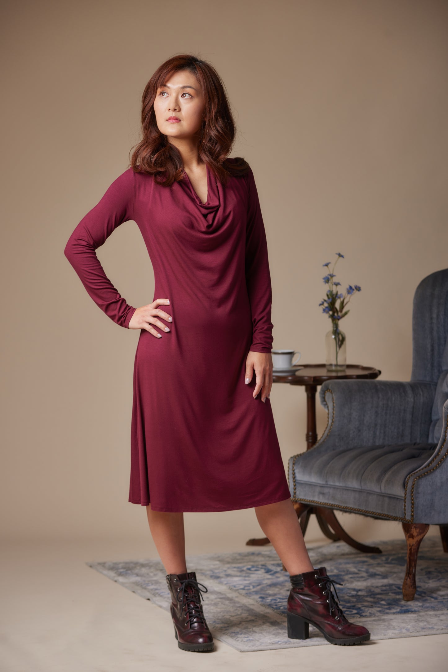Woman wearing a burgundy dress standing in a room with a blue armchair and small table.