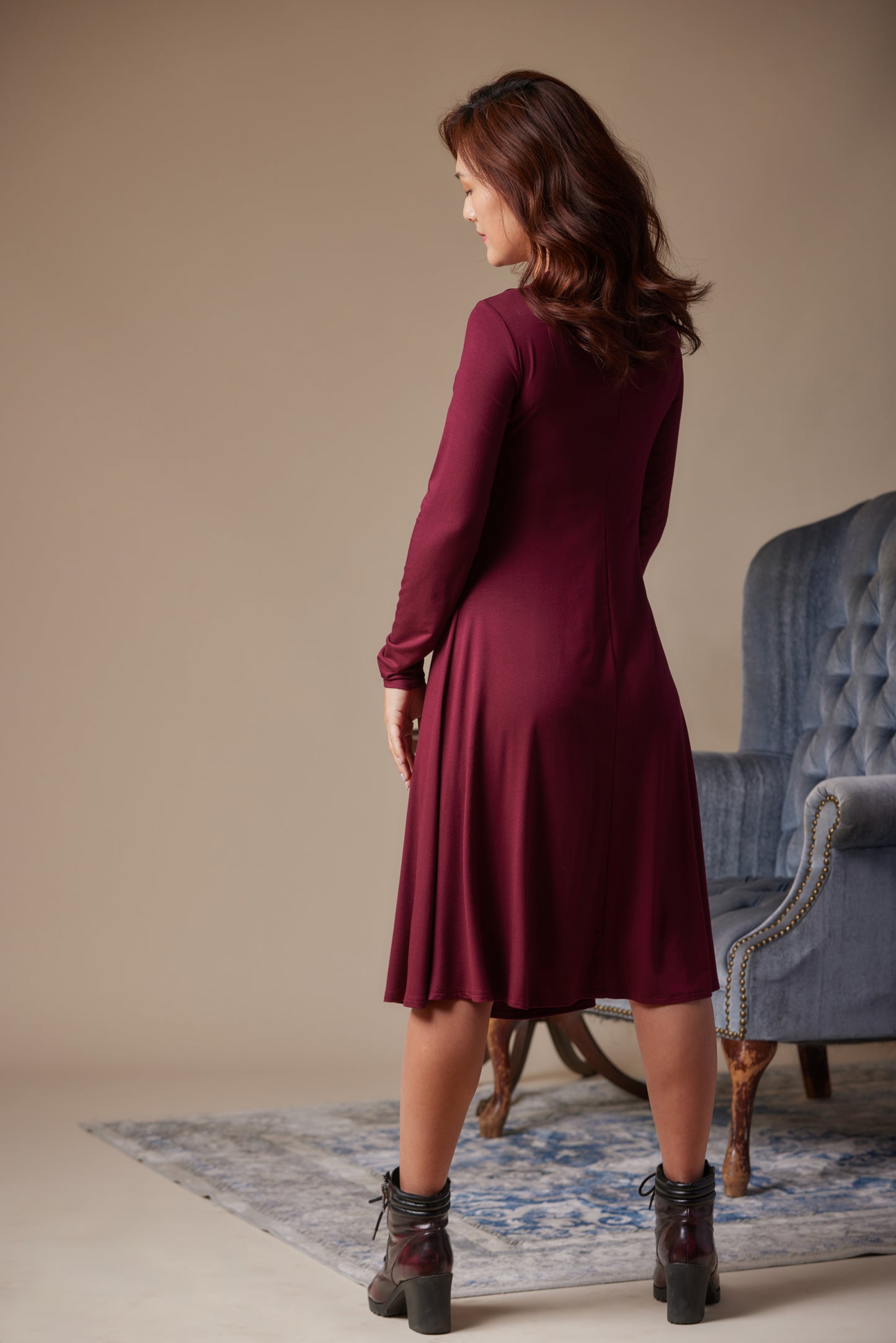 Woman in a burgundy dress standing in a room with a blue armchair and patterned rug.