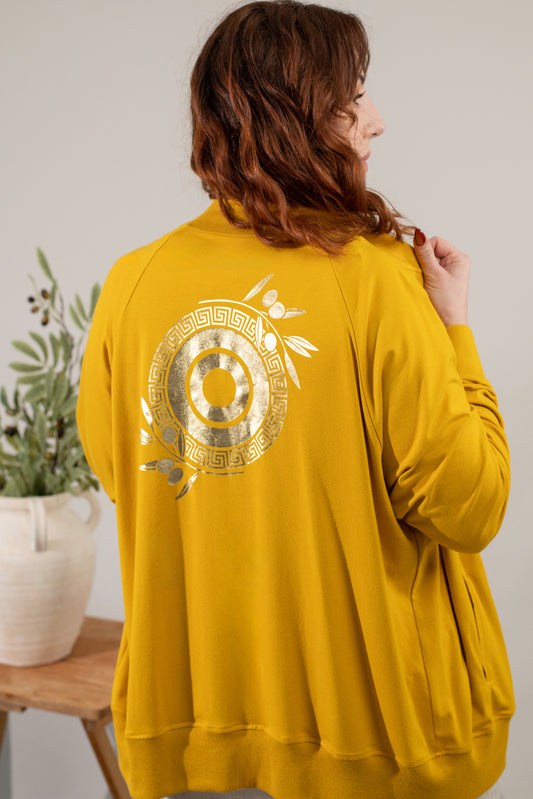 Person wearing a mustard yellow jacket with a gold design on the back, standing in a room with a plant and wooden table.