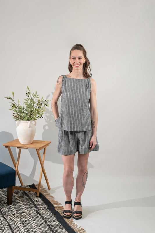 Woman wearing a gray checkered outfit standing in a minimalistic room with a plant and small table.