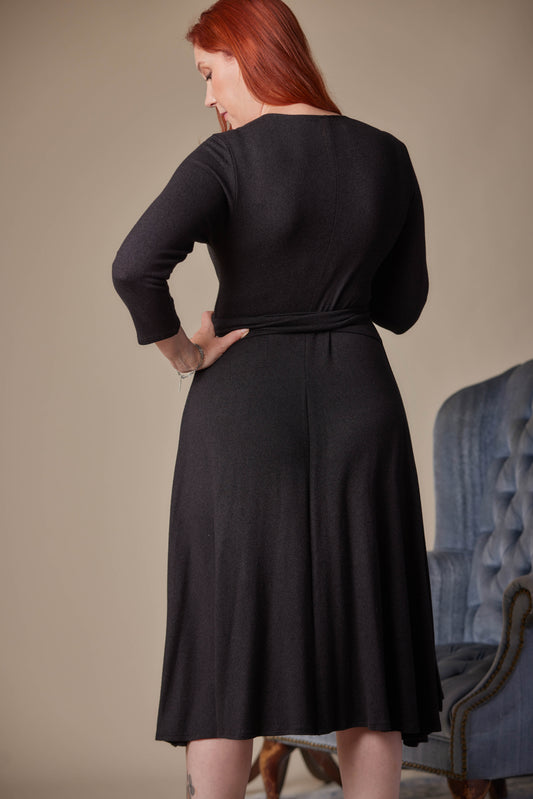 Woman wearing a black dress standing in front of a beige wall with a blue armchair.