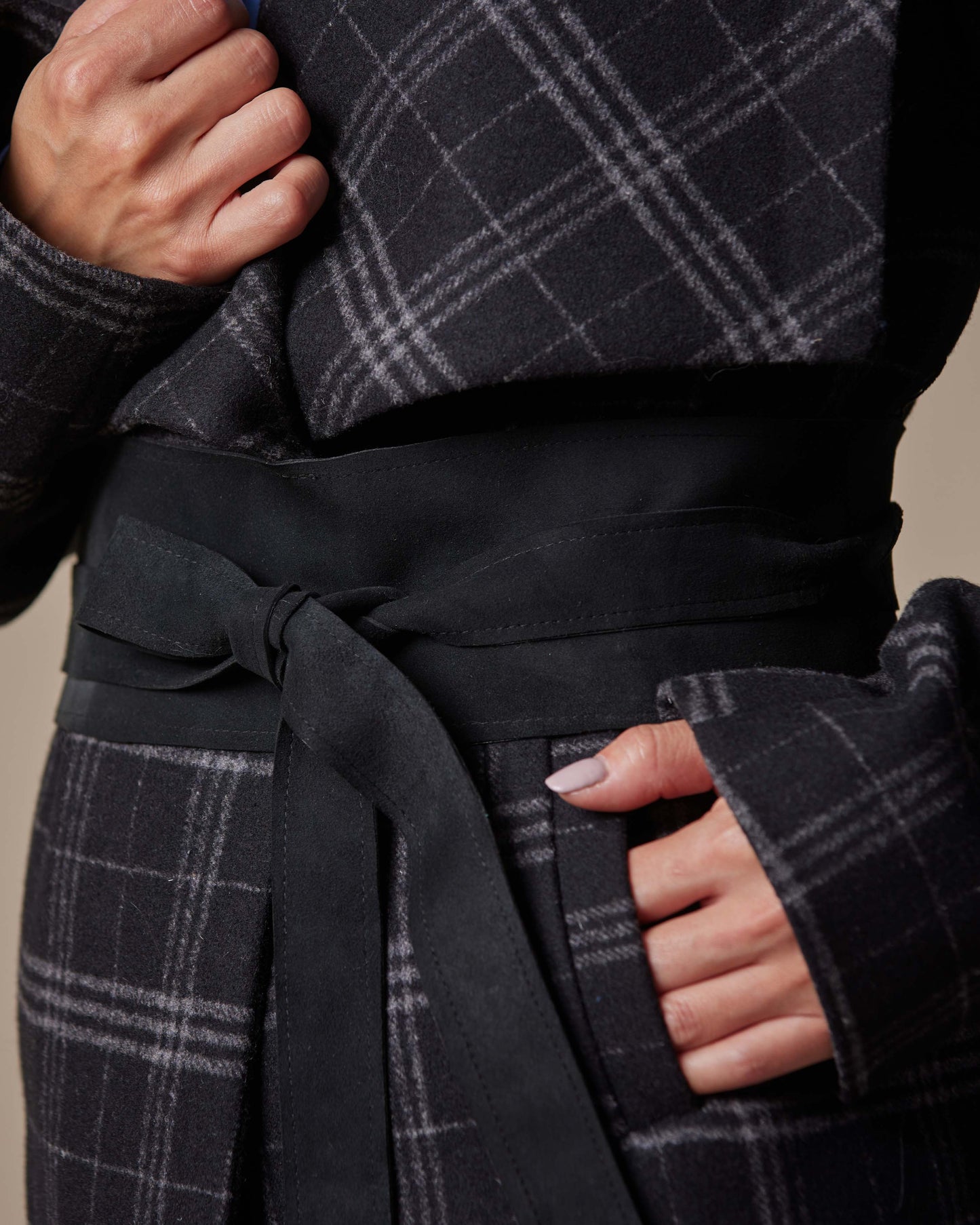 Person wearing a dark gray plaid coat with a belt against a beige background