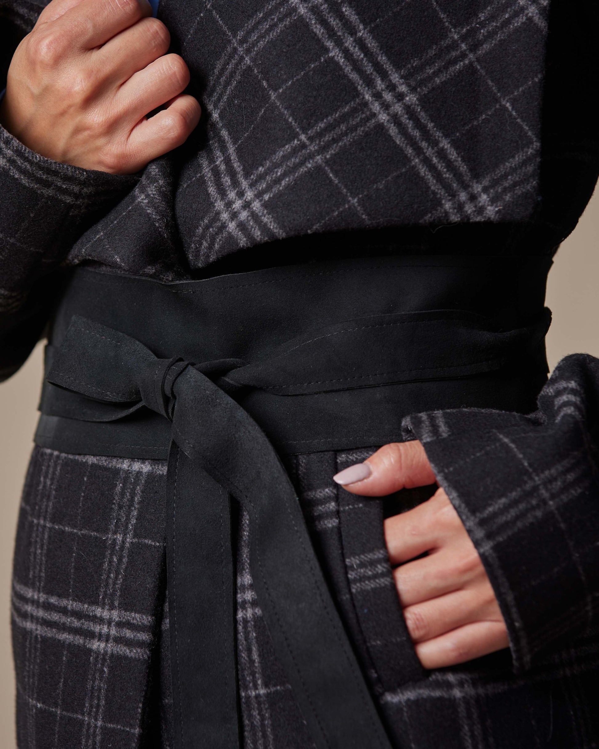 Person wearing a dark gray plaid coat with a belt against a beige background