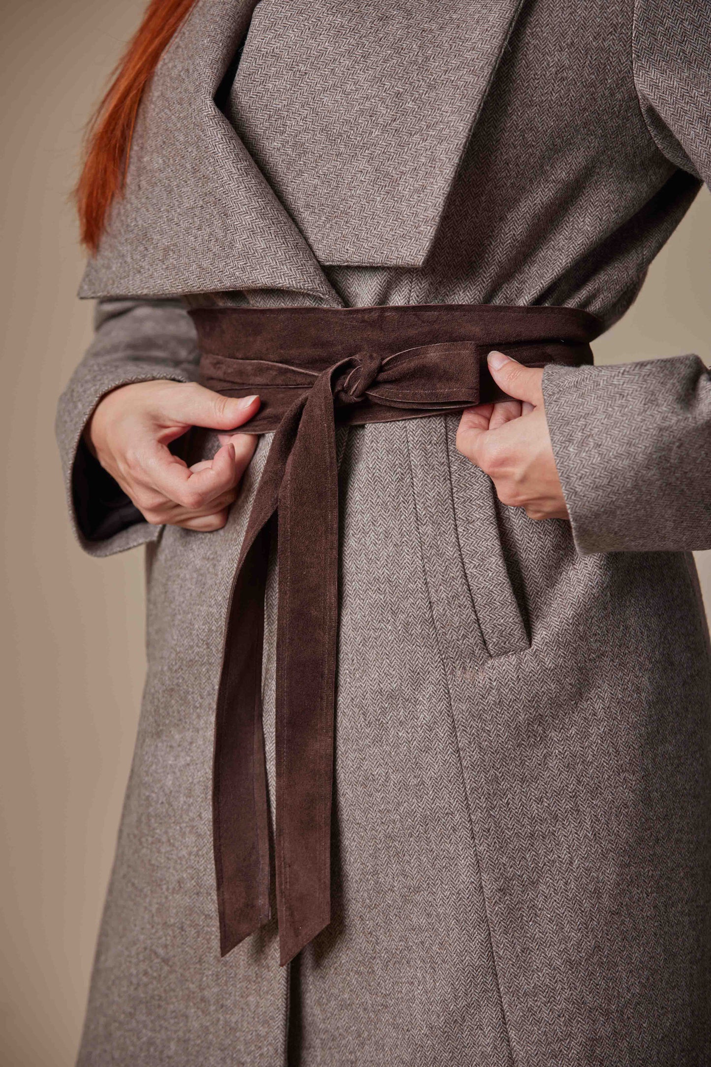Brown coat with a brown belt being adjusted on a neutral background