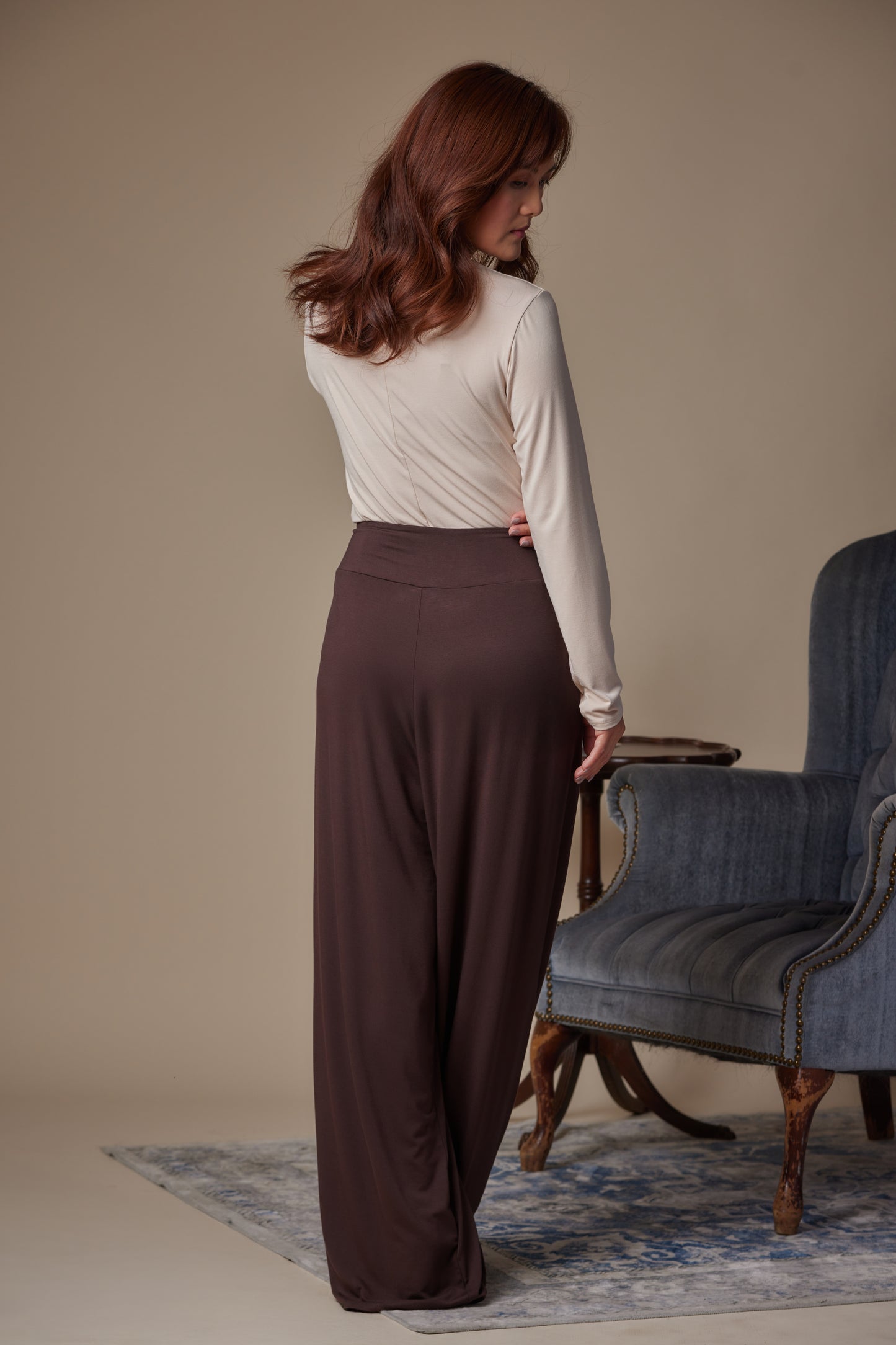 Woman wearing a white long-sleeve top and brown wide-leg pants standing in a room with a blue armchair.