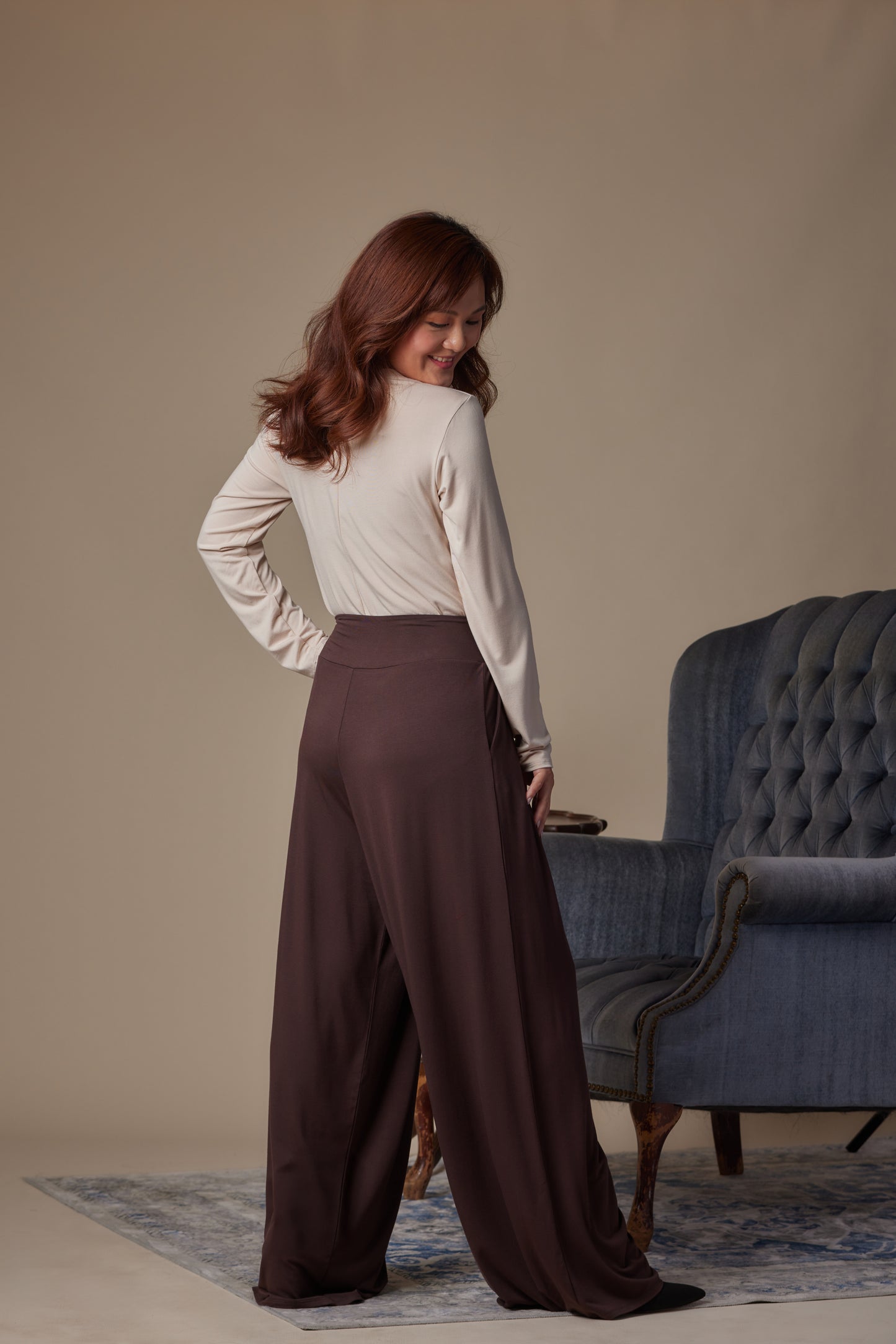 Woman wearing a white blouse and brown wide-leg pants standing in a room with a blue armchair.