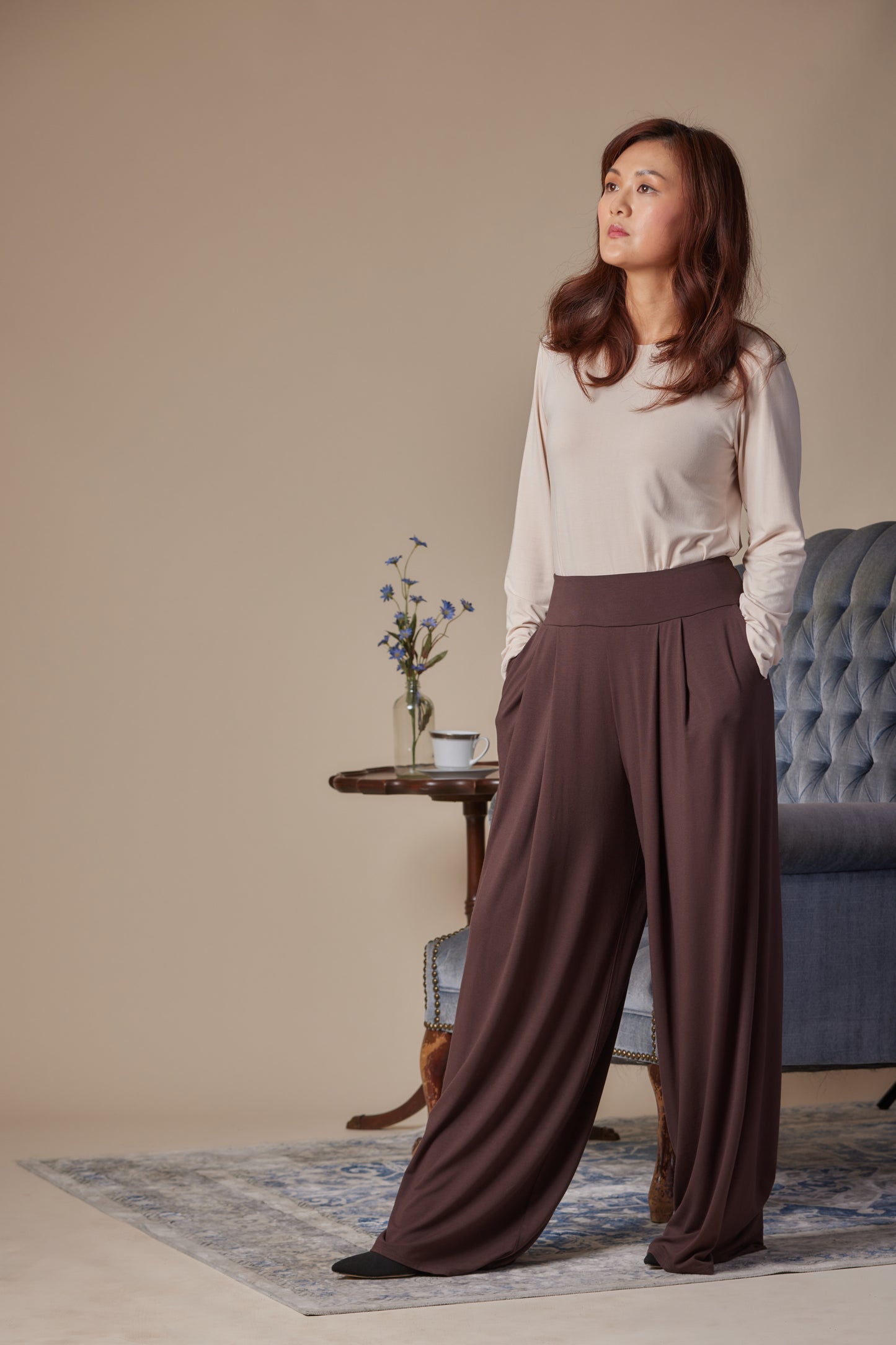 Woman wearing a light blouse and brown wide-leg pants standing in a room with a beige wall and blue sofa.