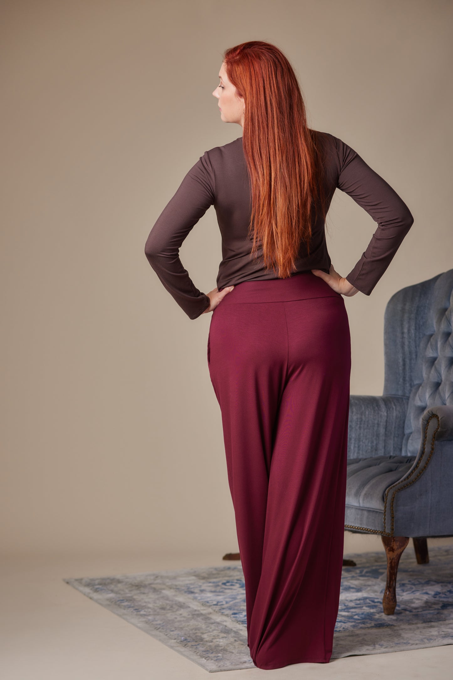 Woman wearing a brown top and burgundy pants standing in a room with a blue chair.