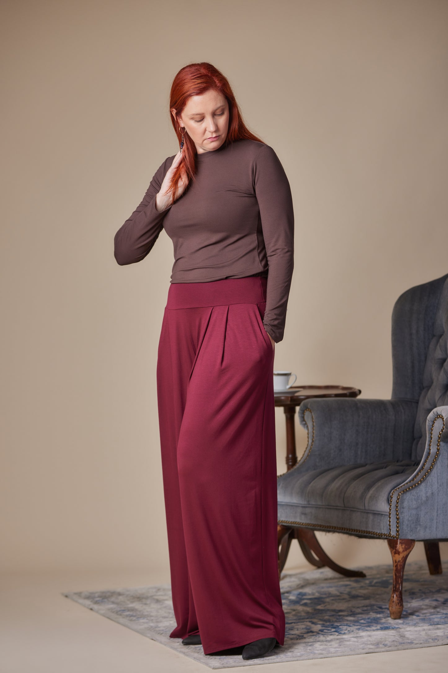 Woman in a brown top and burgundy pants standing in a room with a blue armchair.