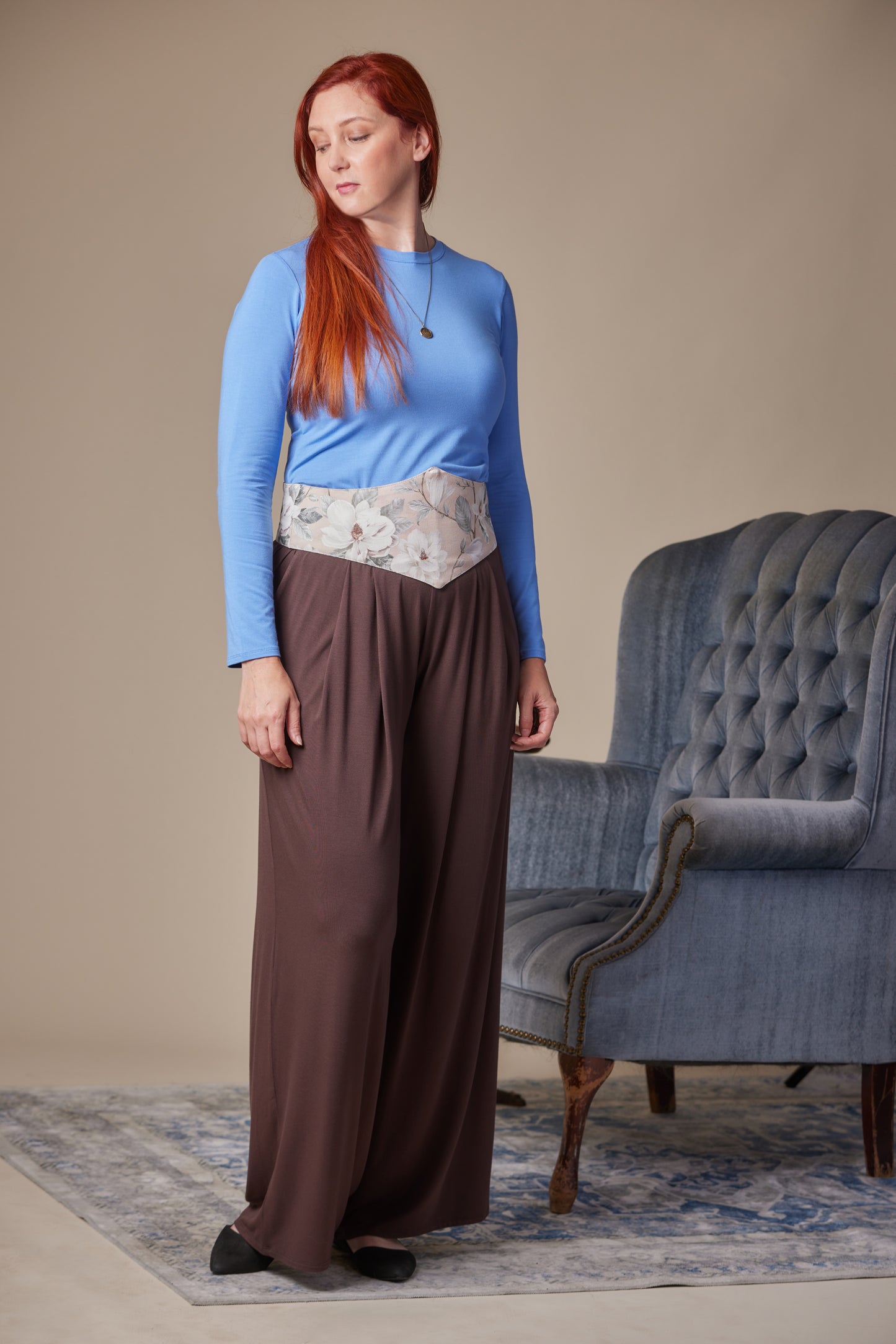 Woman in a blue top and brown pants with floral corset belt standing in a room with a blue armchair.