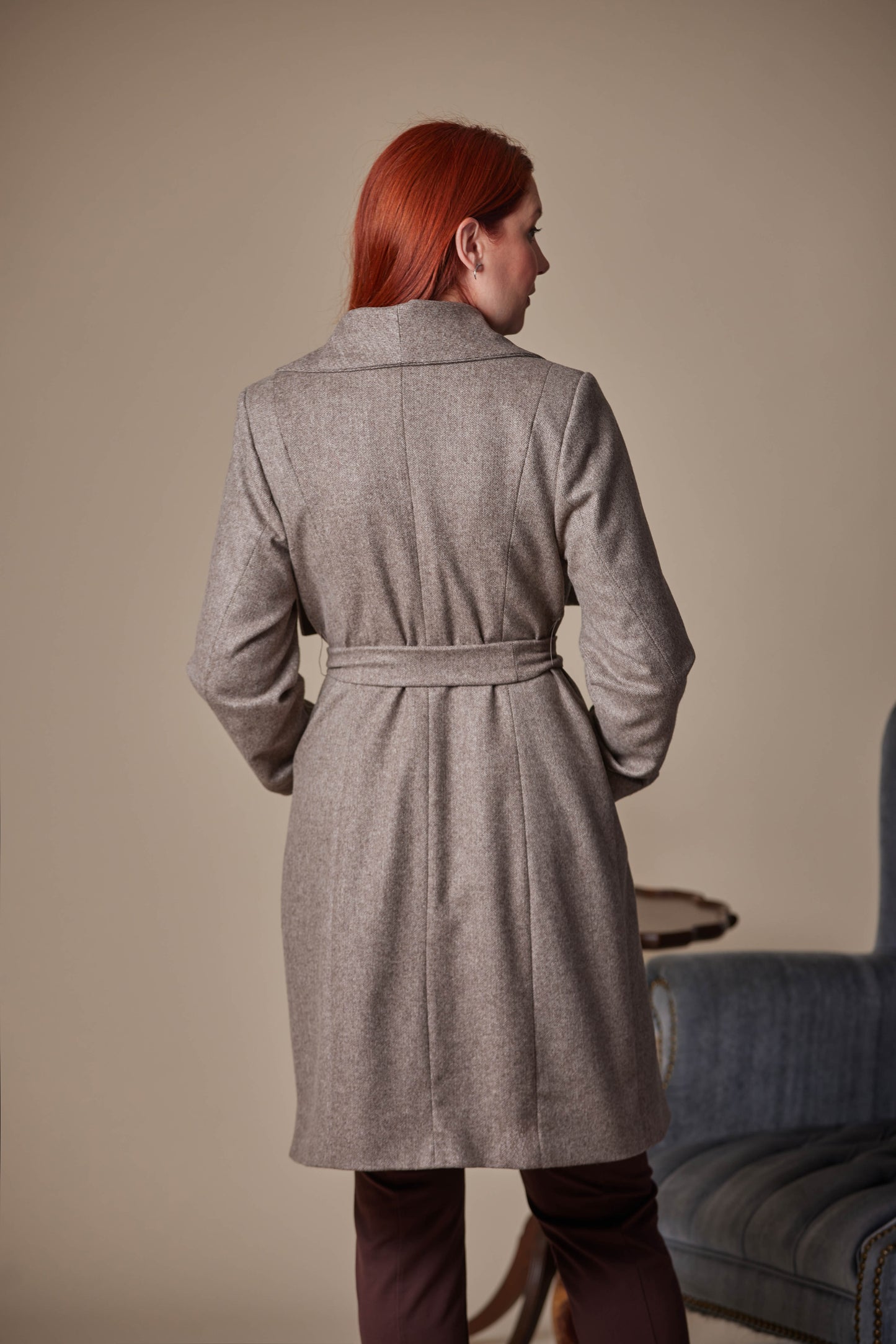 Person wearing a Brown coat standing in a room with a beige wall and a blue chair.