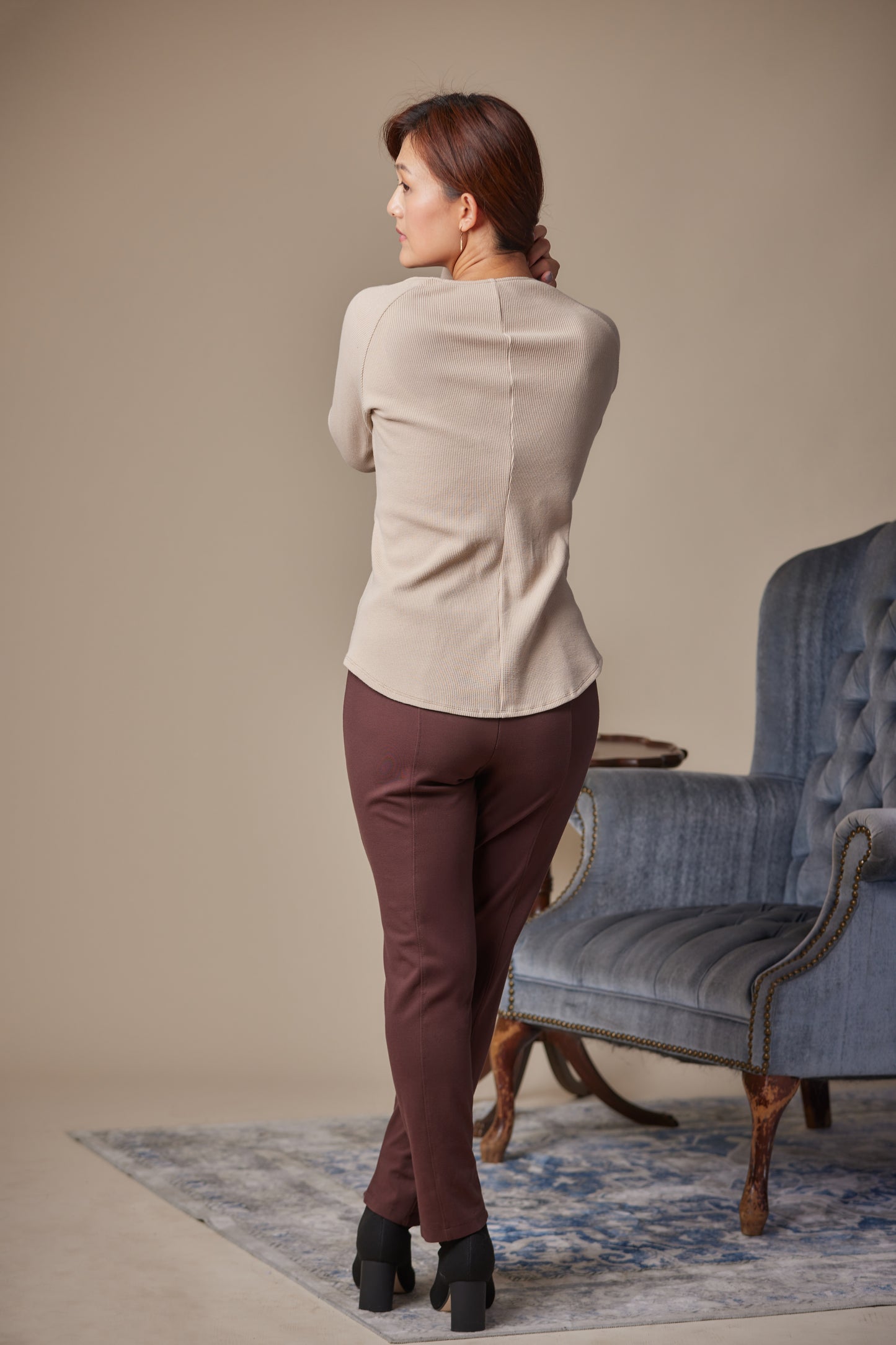 Woman in beige blazer and brown pants standing in a room with a blue armchair.