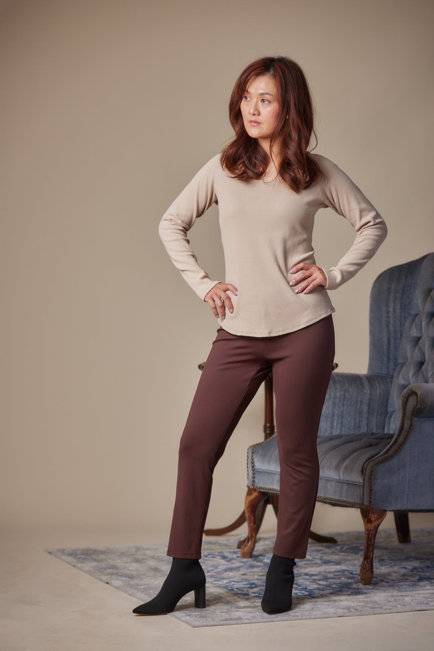 Woman wearing a beige sweater and brown pants standing in a room with a blue chair.