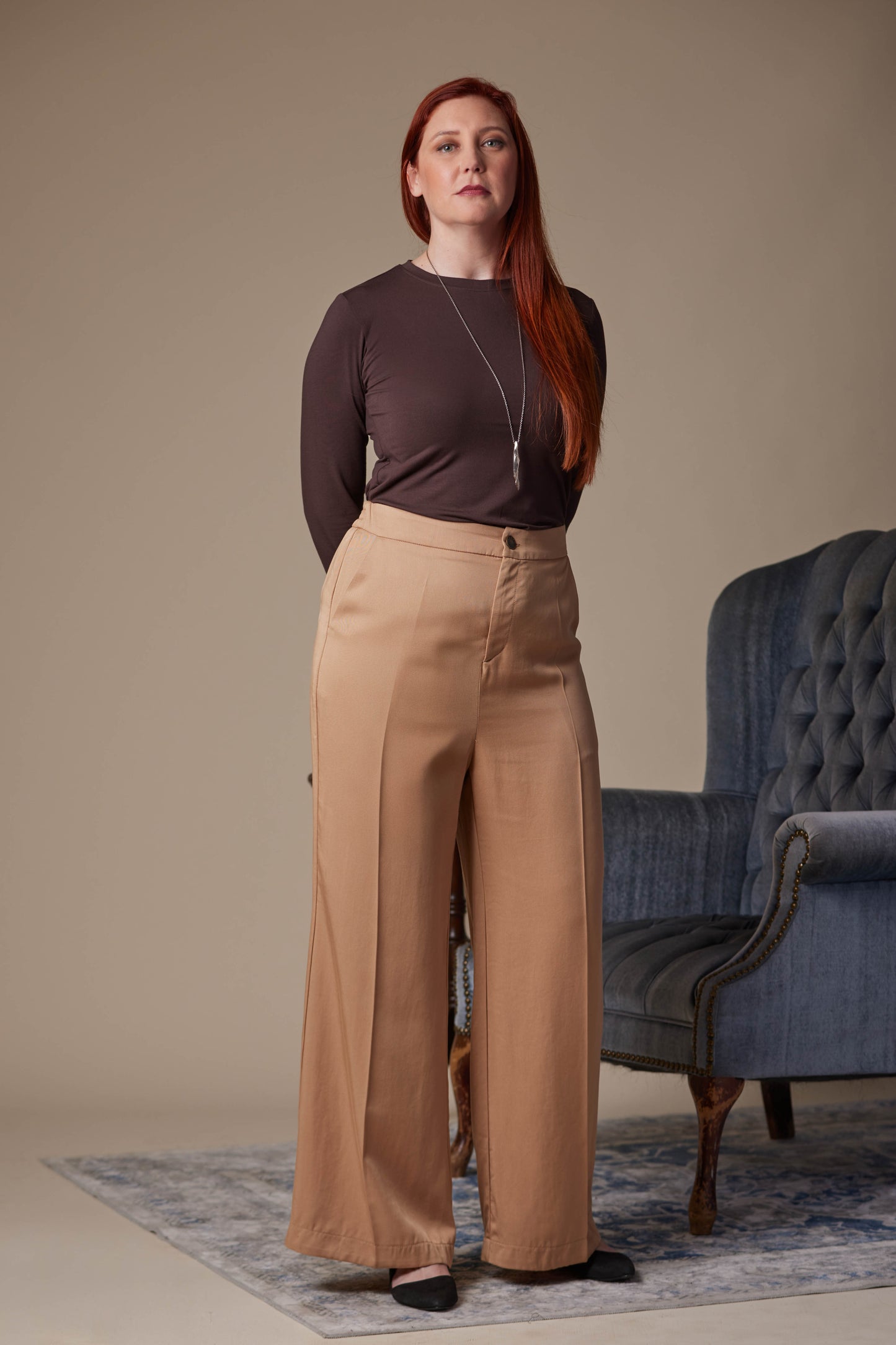 Woman wearing a brown top and beige pants standing in a room with a blue chair.