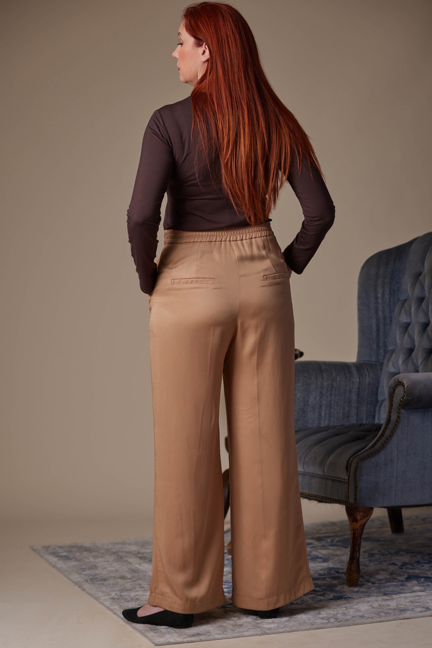 Woman wearing a brown top and beige pants standing in a room with a blue armchair.