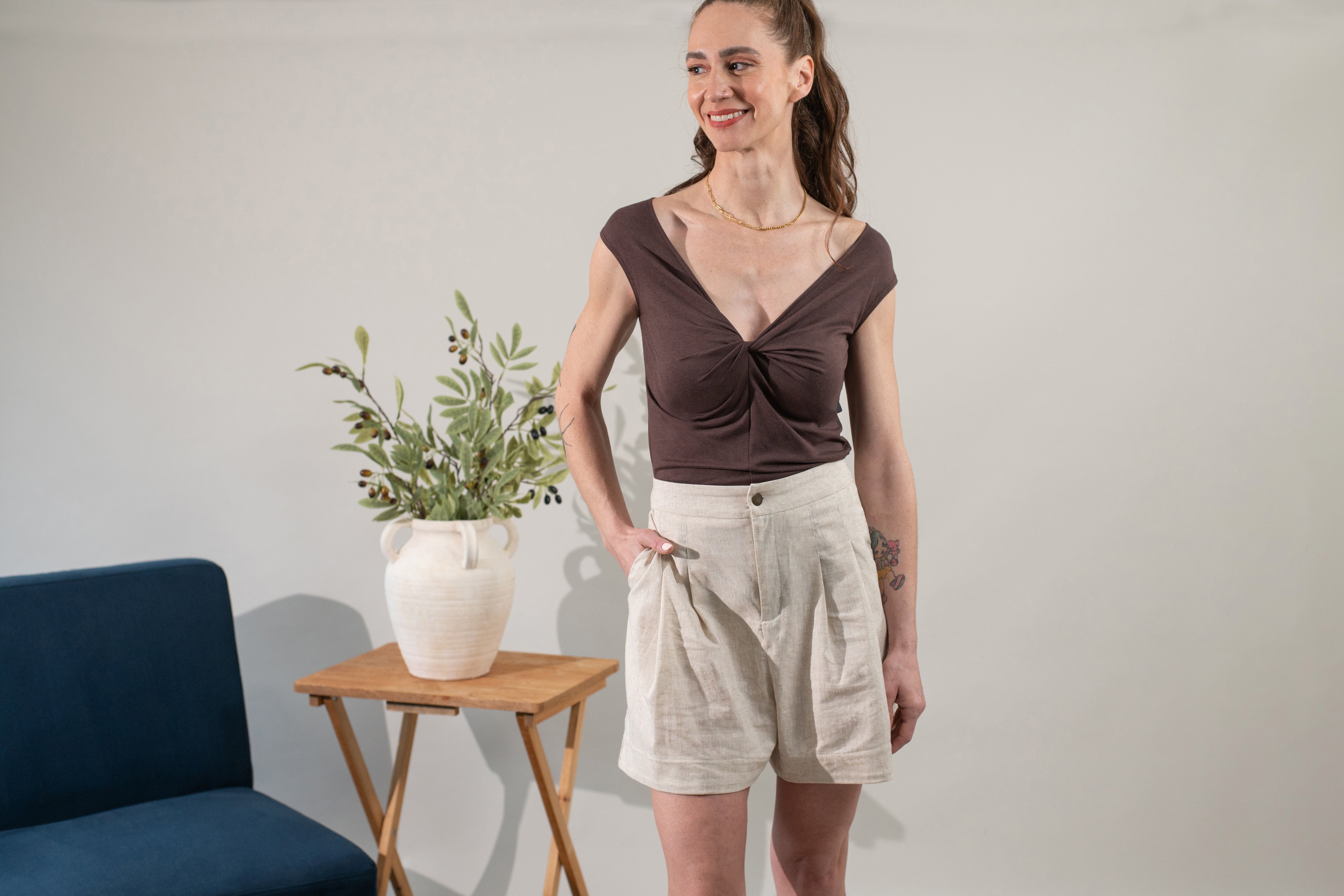 Woman wearing a brown top and beige shorts standing in a room with a plant and table.