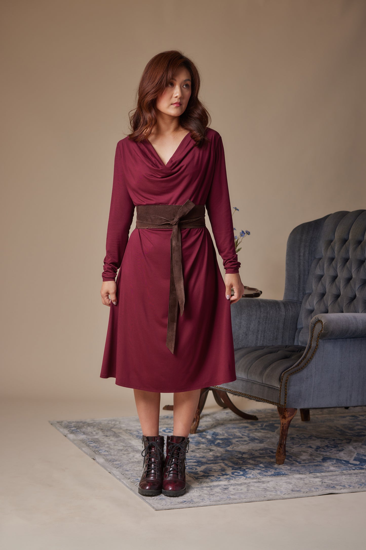 Woman wearing a burgundy dress with a brown belt in a room with a gray armchair.