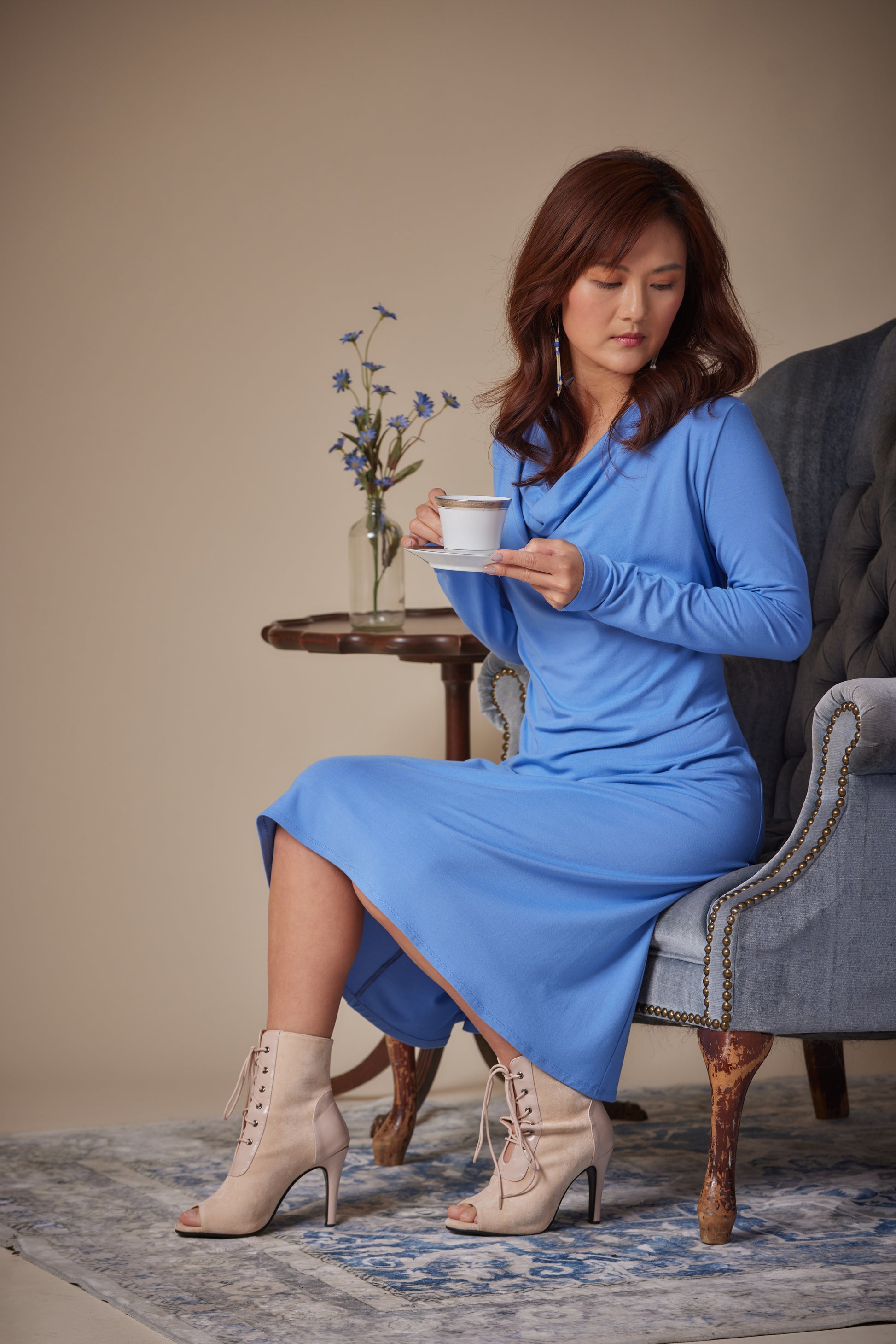 Woman in a blue dress sitting on a chair holding a cup, with a small table and flowers in the background.