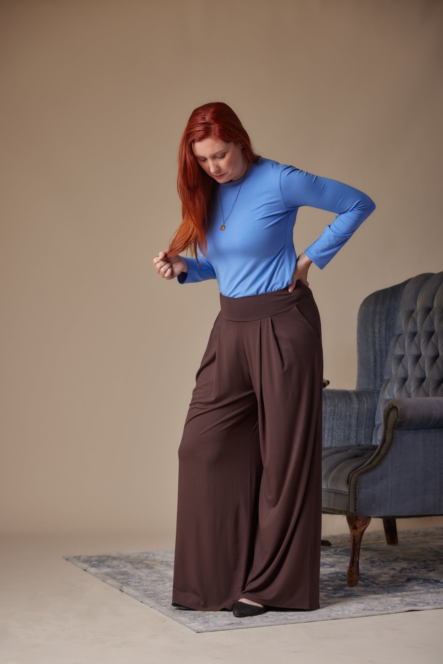 Woman in blue top and brown pants posing in a room with a gray armchair.