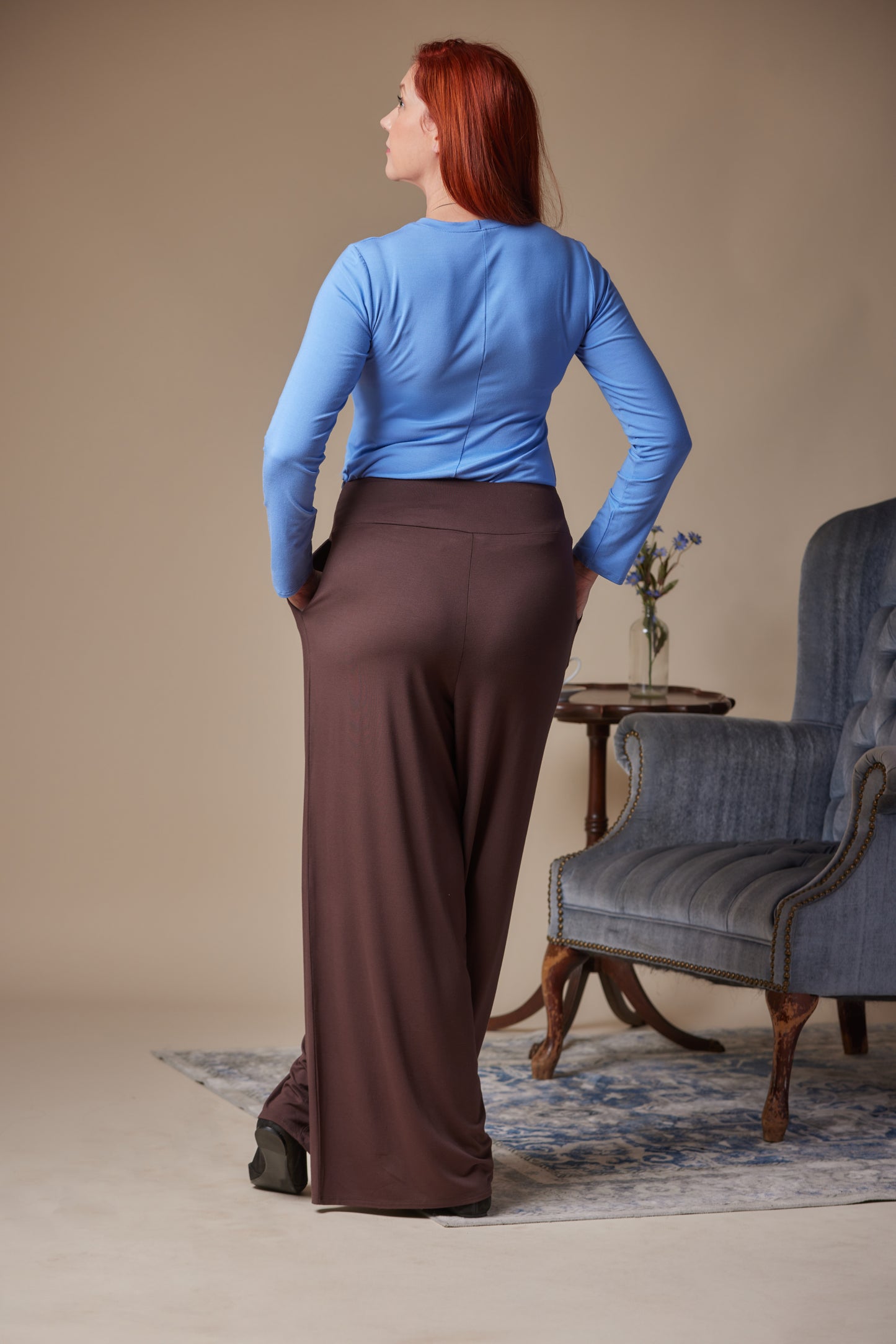 Woman wearing a blue top and brown pants standing in a room with a chair and table.