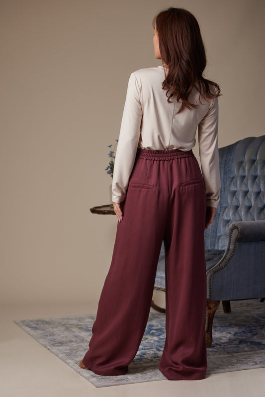 Woman wearing a white blouse and burgundy pants standing in a room with a blue chair and beige wall.