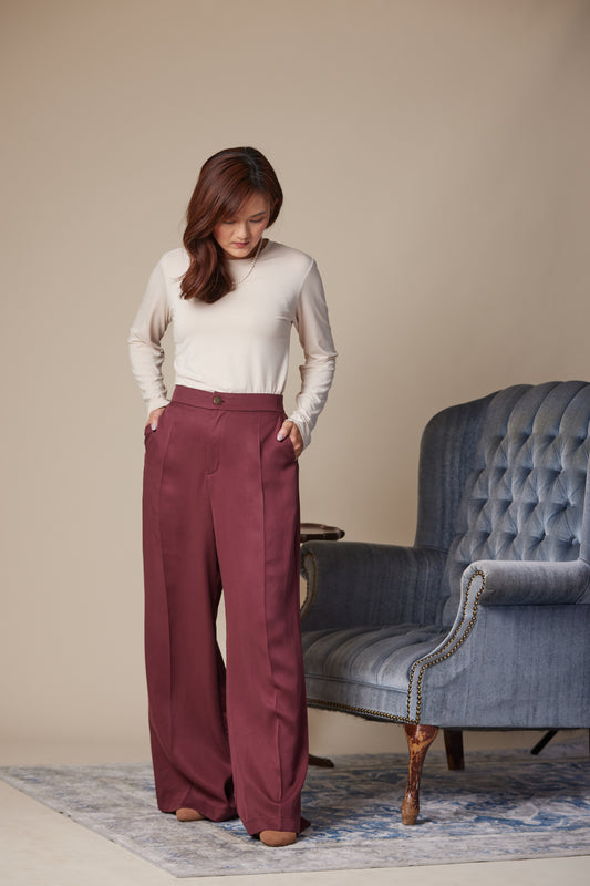 Woman wearing a white top and maroon pants standing in a room with a blue upholstered chair.