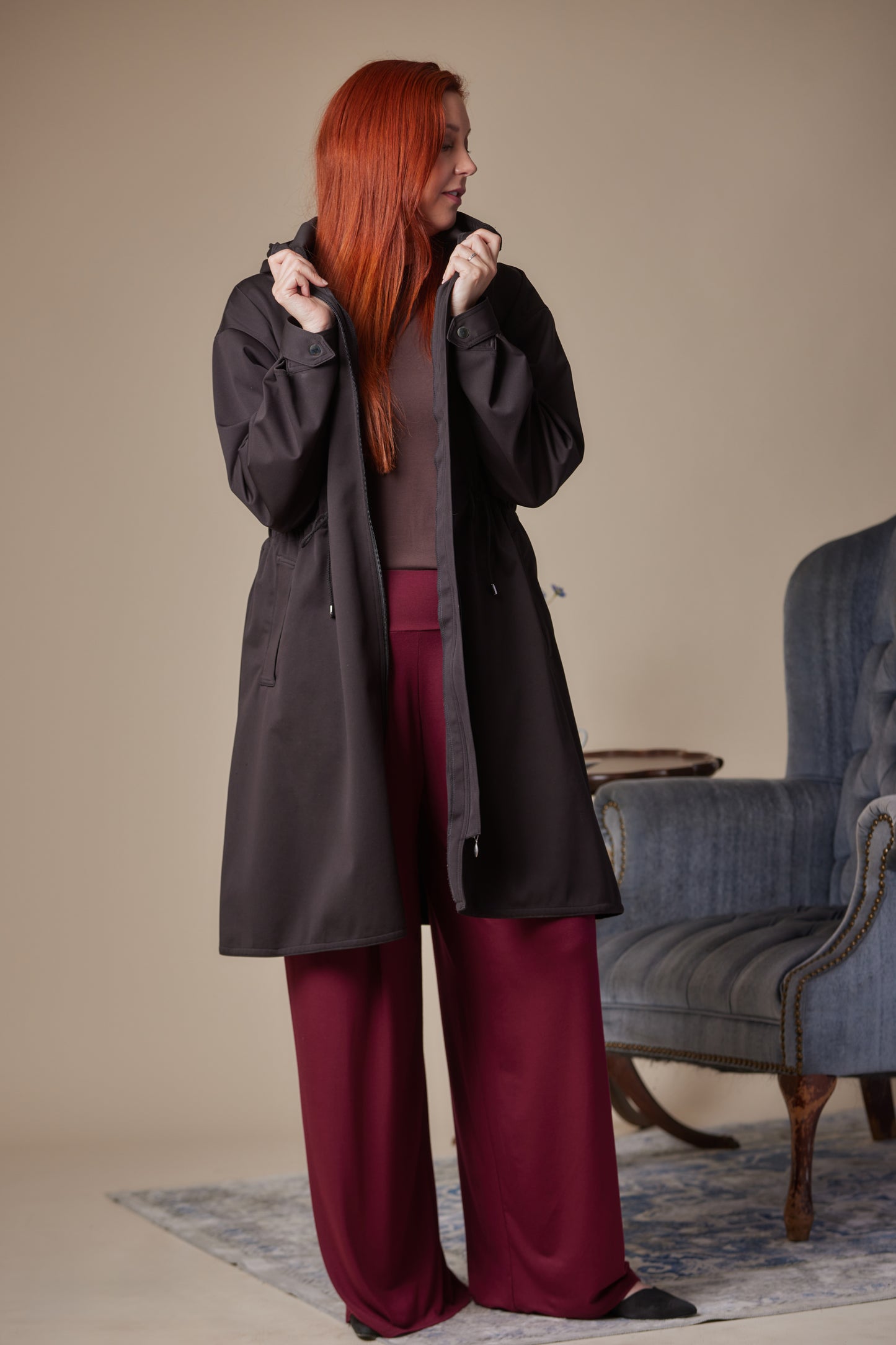 Person wearing a long black coat and burgundy pants standing in a room with a blue armchair.