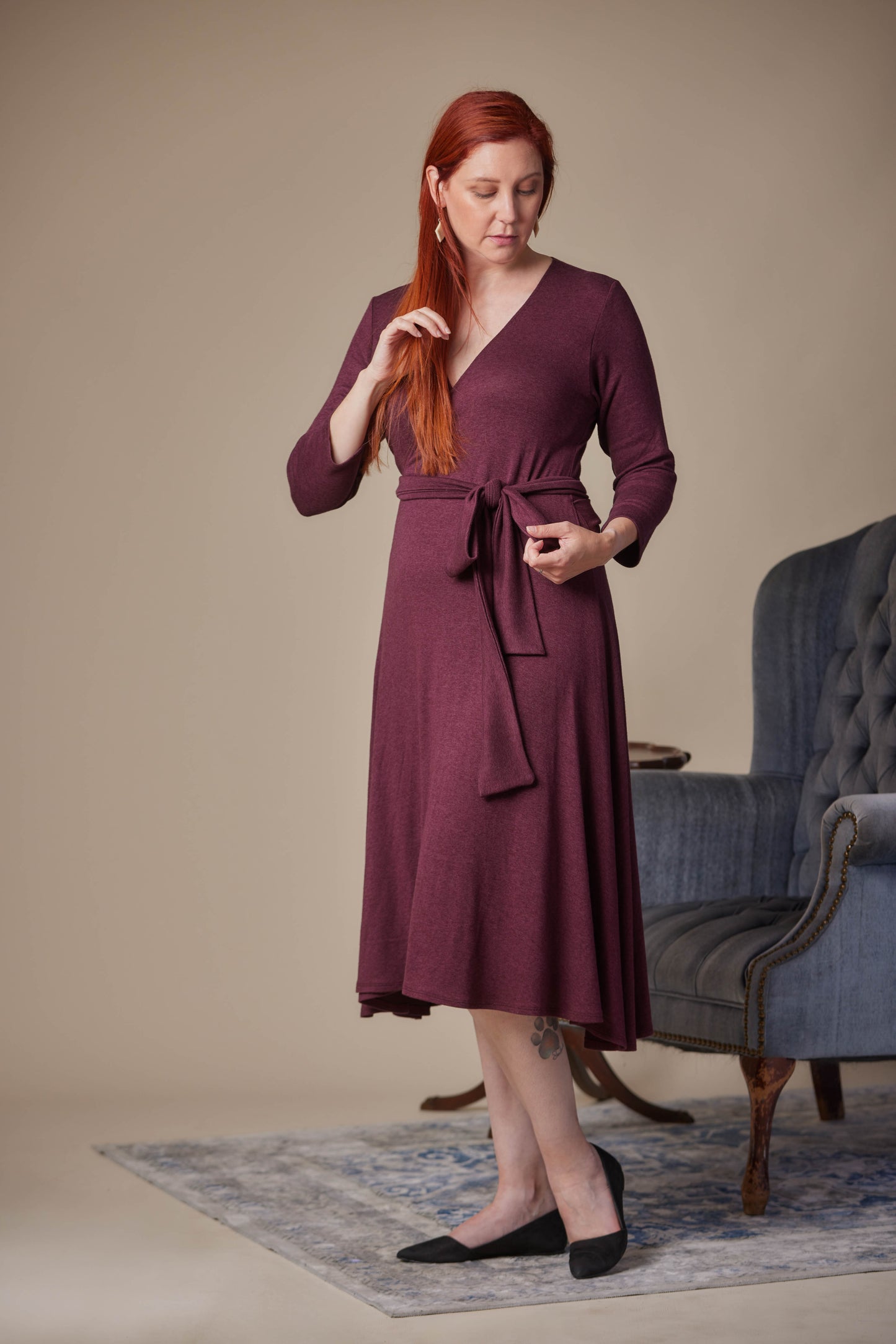 Woman wearing a burgundy dress standing in a room with a blue armchair.