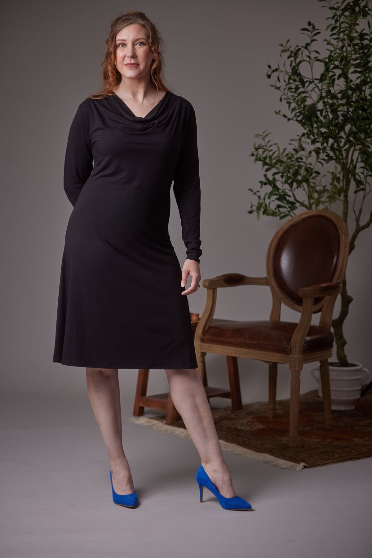 A pregnant woman modeling a black long-sleeve cowl-neck dress, standing in a studio with a gray background, a wooden armchair, and a potted plant. She is wearing bright blue high heels.