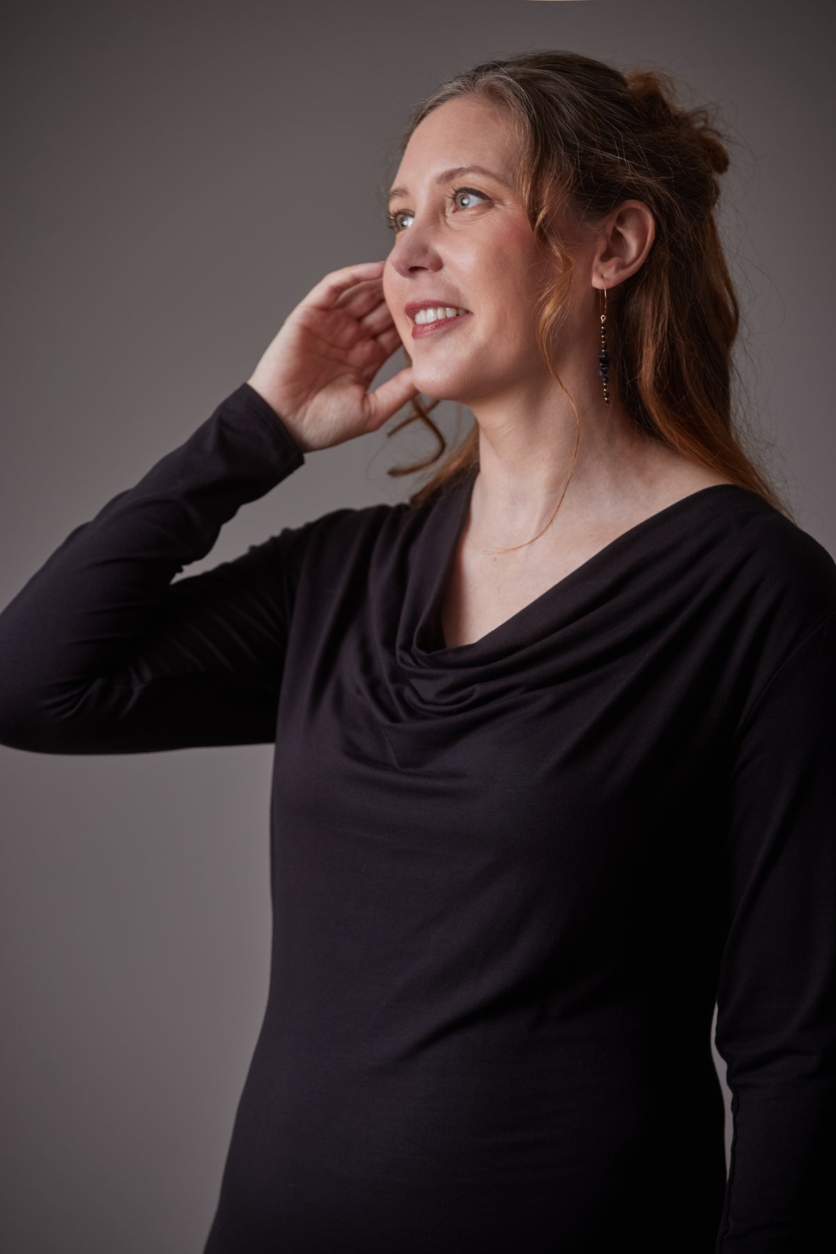 A woman with light brown hair in a half-up bun wearing a black long-sleeve cowl neck dress, posing against a dark gray studio background.