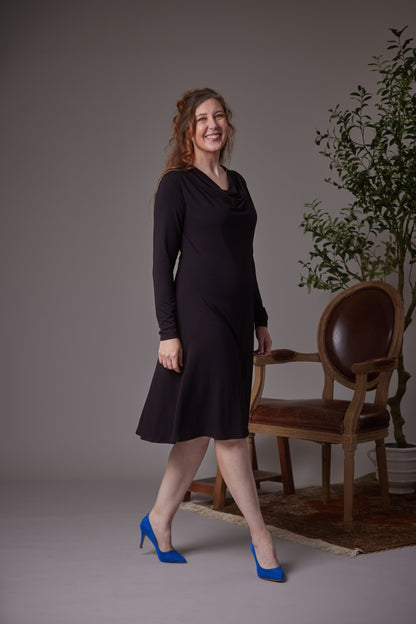 A woman modeling a black long-sleeve cowl-neck midi dress, standing in a studio setting next to a brown leather and wood armchair with a potted plant in the background.