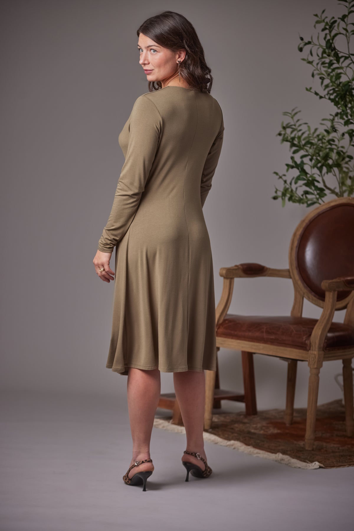 A woman modeling a long-sleeve, knee-length fern dress, shown from a three-quarters back view in a studio setting with a wooden chair and plant in the background.