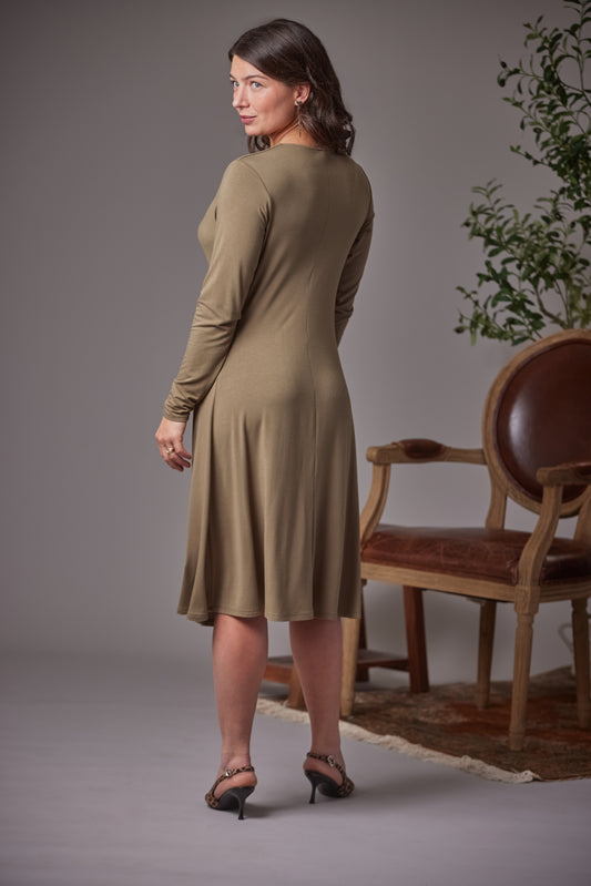 A woman modeling a long-sleeve, knee-length fern dress, shown from a three-quarters back view in a studio setting with a wooden chair and plant in the background.