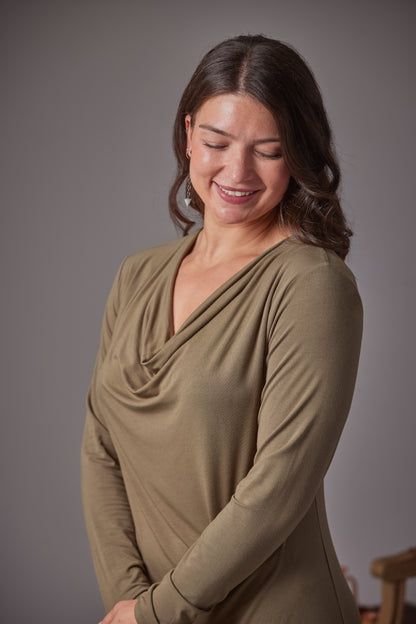 A woman with dark brown hair is wearing a long-sleeve, cowl-neck top in a muted fern color, smiling and looking down against a solid gray studio background.