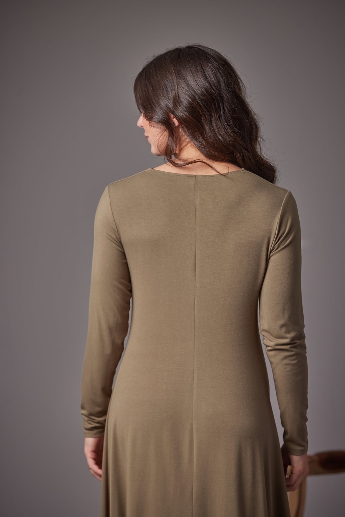 A woman viewed from the back wearing a long-sleeve, knee-length knit dress in a muted fern color against a solid gray studio background.